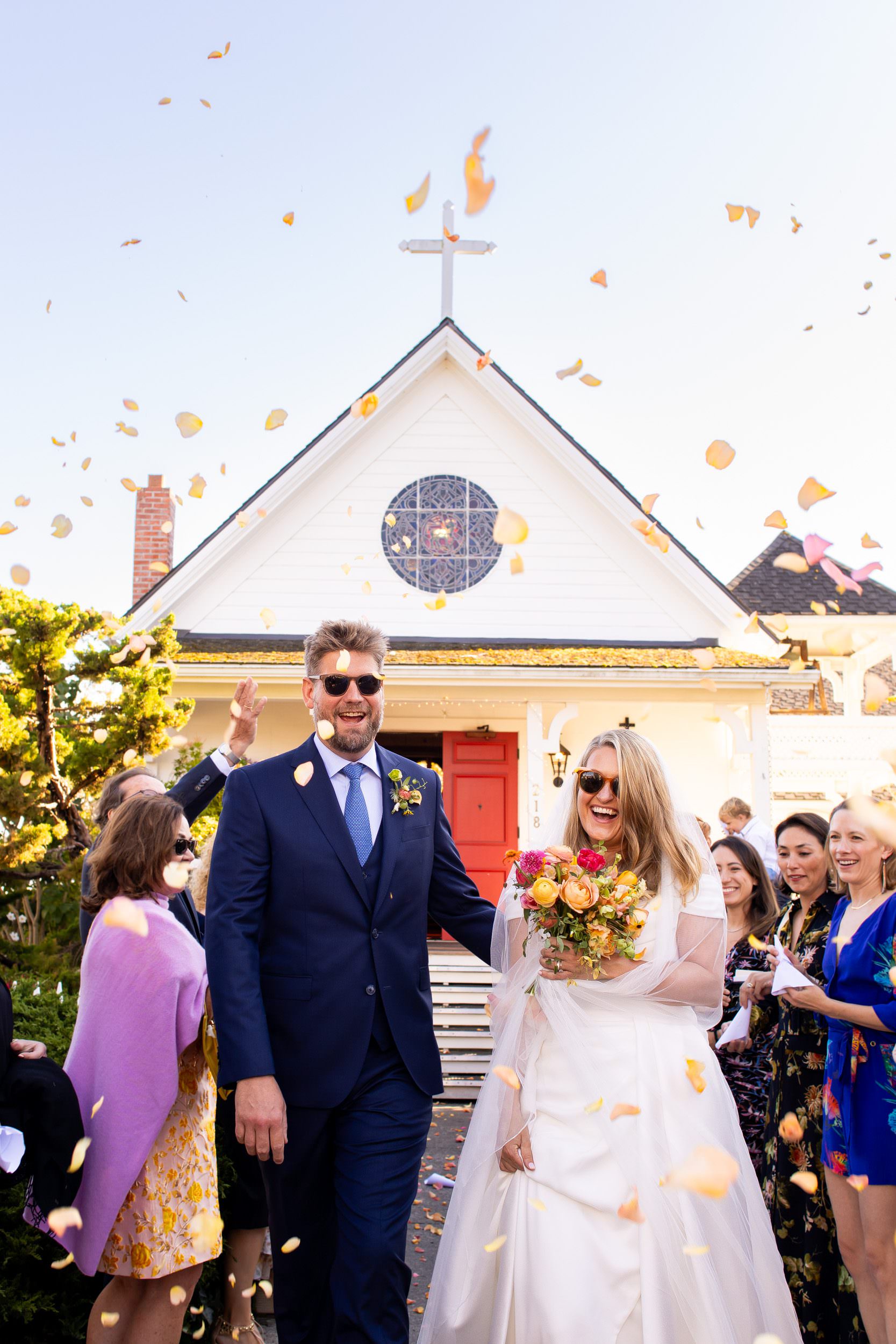 Bride and groom exiting wedding reception in the Eastsound of Orcas Island at the Orcas Island Community Church wearing sunglasses as their guests throw rose petals