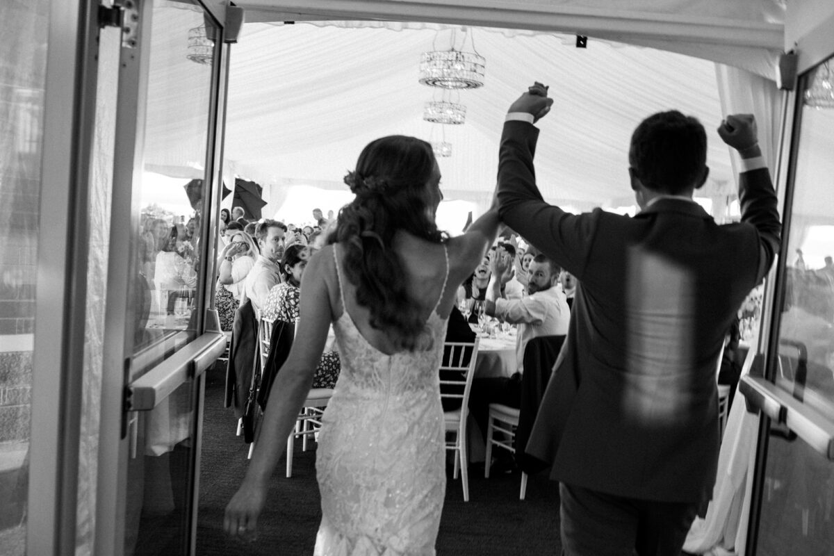 Black and white photo from behind a bride and groom, entering Woodmark hotel wedding reception with guests cheering