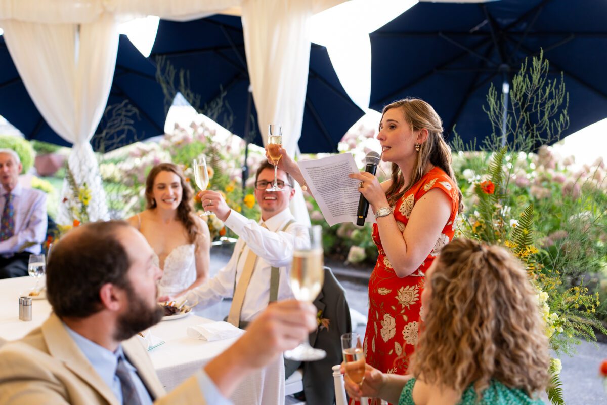 Sister of the groom, giving a speech and raising her glass during speeches under the tent for the wedding reception