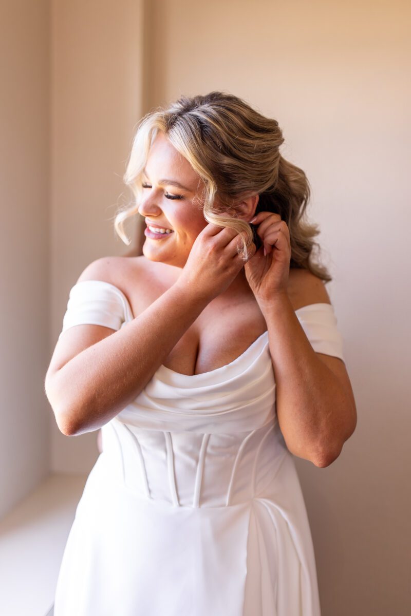 Bride smiling while she puts in her earrings at the Hotel Leo ahead of her Bellingham Washington wedding. Wedding dress with boning, strapless off the shoulder wedding dress